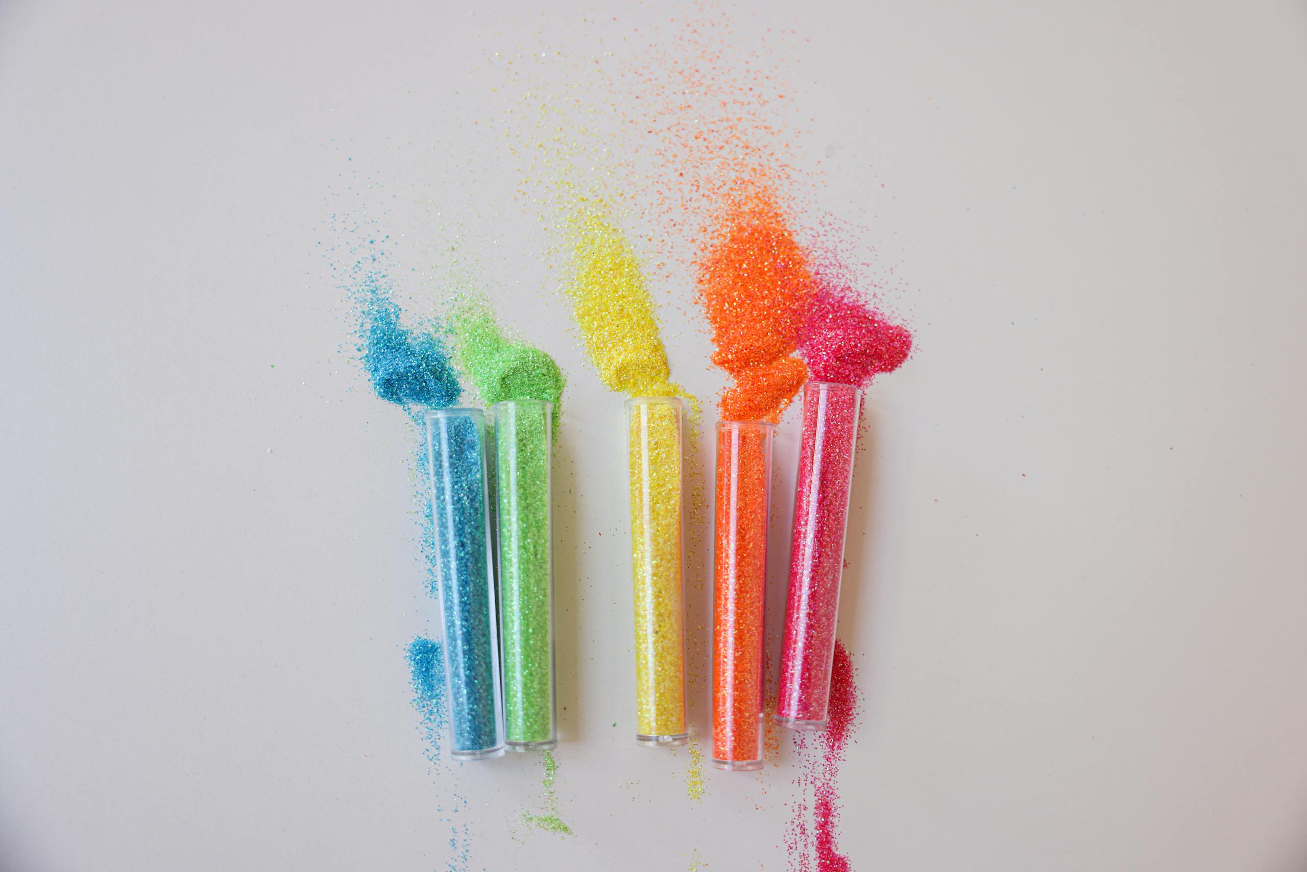Four tubes containing colorful glitter powders spilled on a white surface, creating a vibrant display.